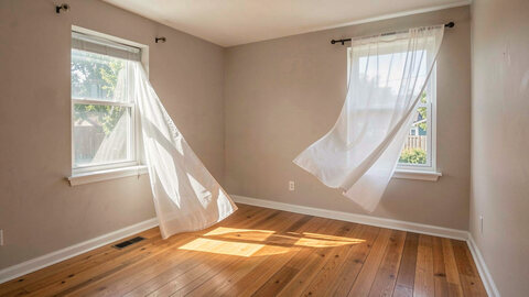 Two windows open on opposite walls of a small room, a light curtain lifting in the draught and daylight crossing the floor.